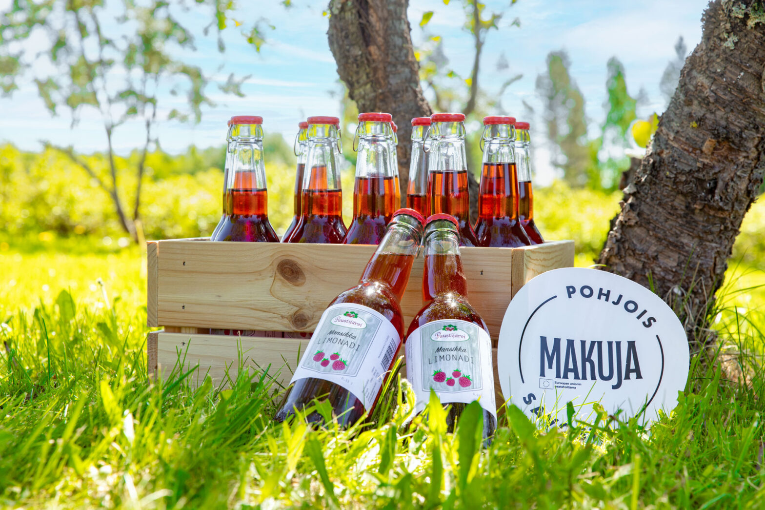 Puustijärven marjatilan mansikkalimonadi pulloja puisessa korissa vihreälle nurmelle aseteltuna.