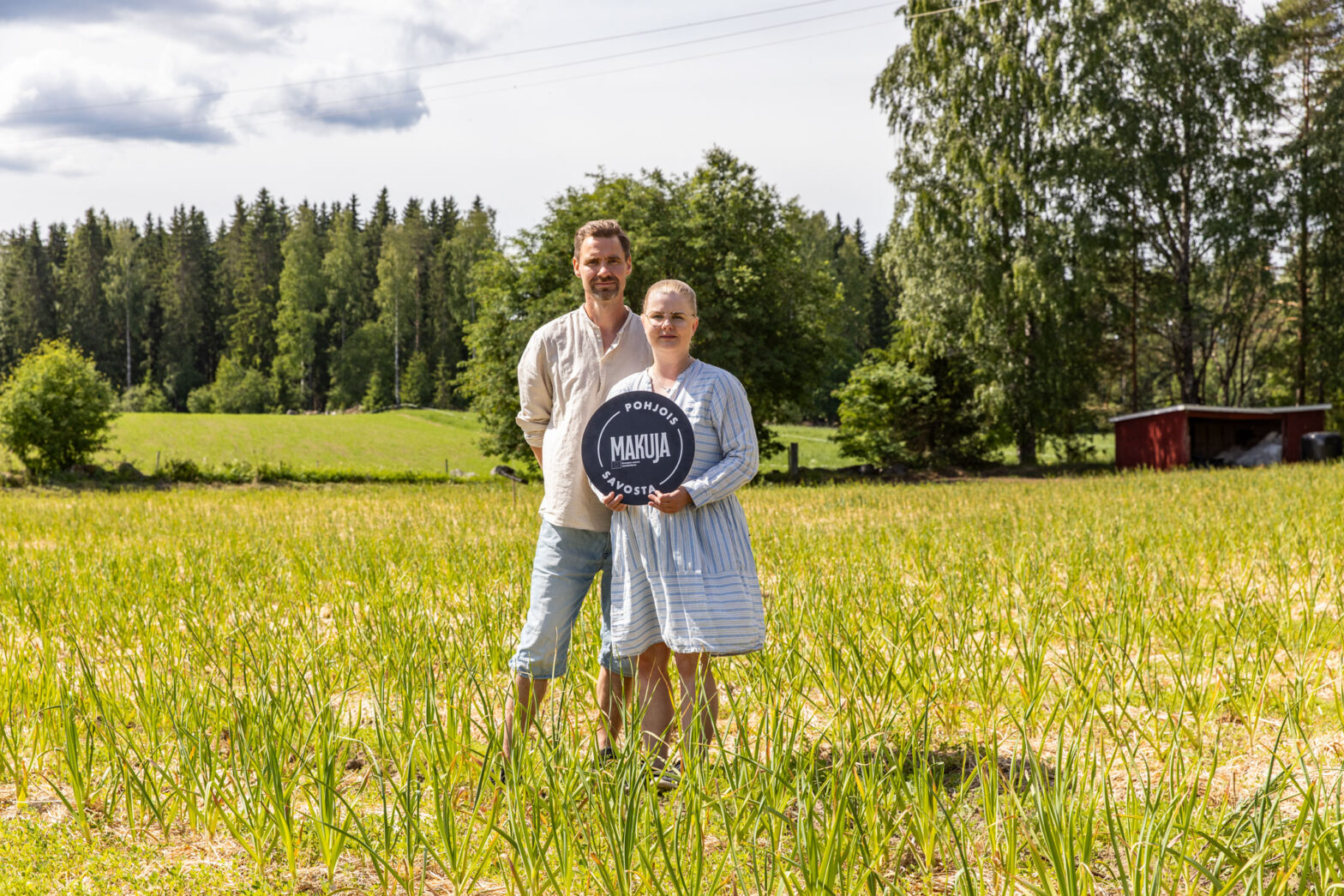 Kankaanrinteen tilan yrittäjäpariskunta seisoo keskellä valkosipulipeltoa pidellen makuja Pohjois-Savosta kylttiä.